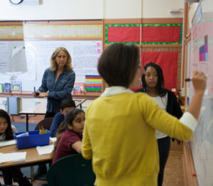 A teacher with a clipboard observes a teacher and students working on a white board as part of a research lesson