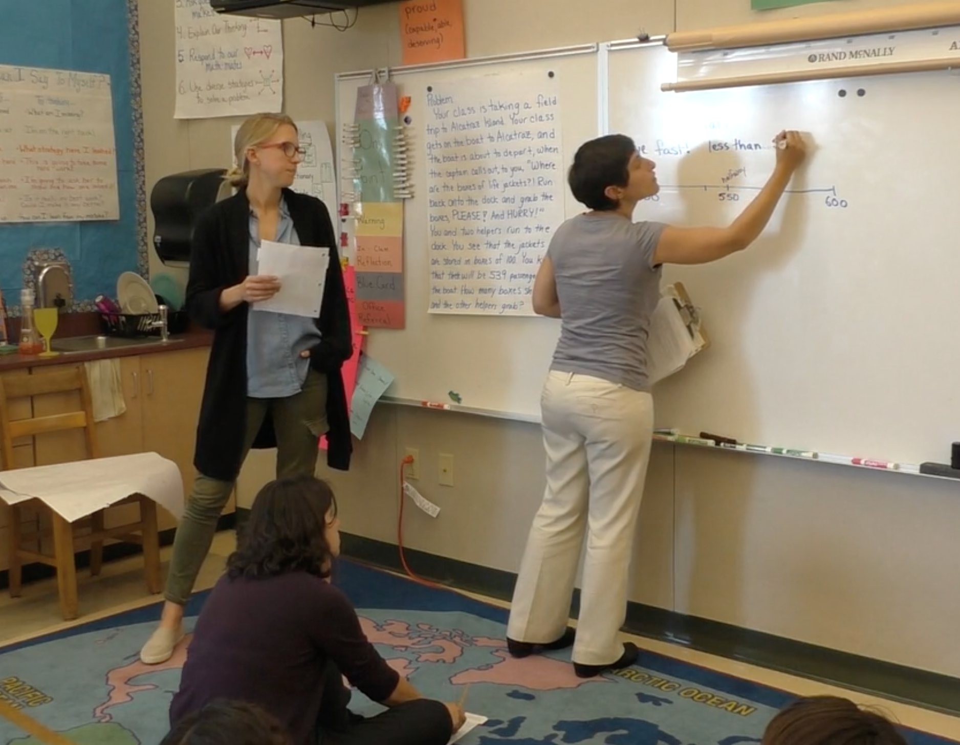 Teachers do a run-through of a lesson, with one teacher writing on a white board, another sitting on a carpet playing the student role, and a third observing