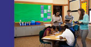 Three teachers write observations on clipboards as they watch four students sitting at a table work on a project
